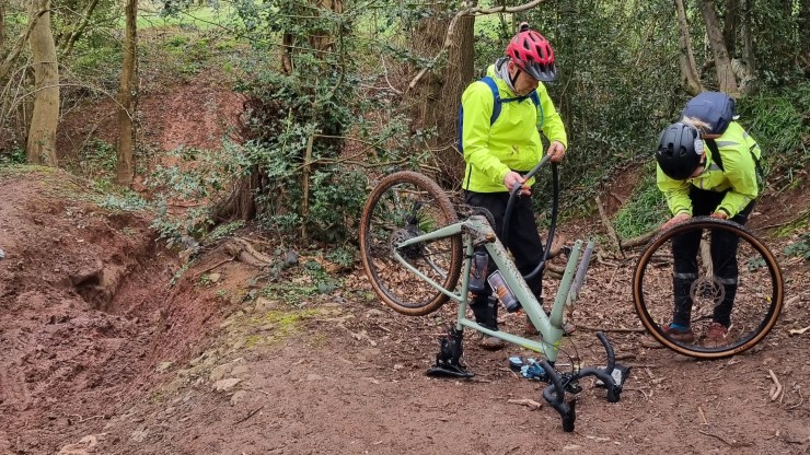 Two cyclists wearing bright yellow jackets and helmets performing maintenance on a bicycle with a removed front wheel in a wooded area.