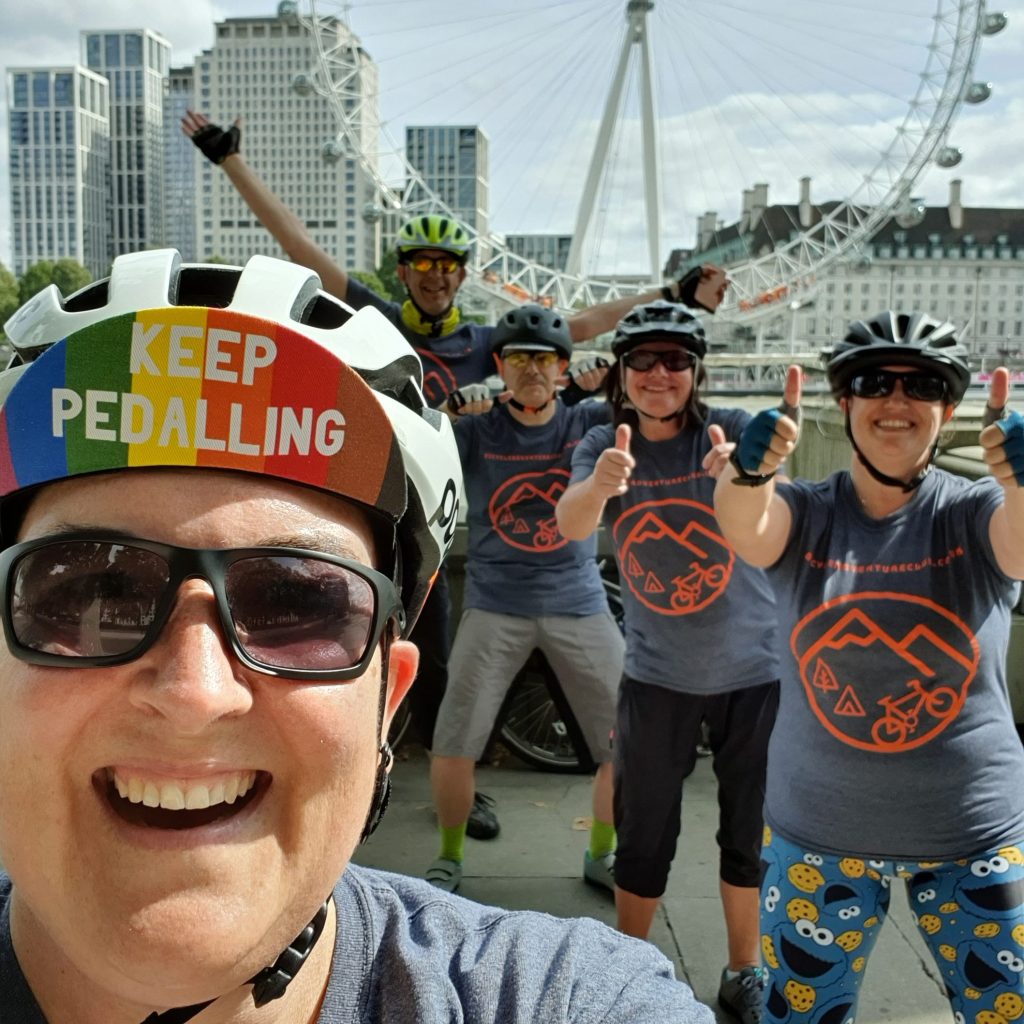 A group of six cyclists posing for a selfie in front of the London Eye, all wearing helmets and matching t-shirts, with one cyclist holding a sign that says 'KEEP PEDALLING'. The mood is cheerful and energetic.